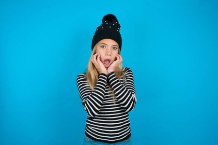 Photo for Stupefied beautiful kid girl wearing knitted black hat and striped turtleneck over blue background expresses excitement and thrill, keeps jaw dropped, hands on cheeks, has eyes popped out - Royalty Free Image