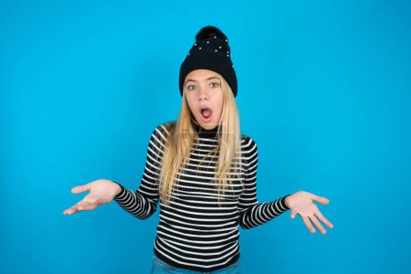 Frustrated beautiful kid girl wearing knitted black hat and striped turtleneck over blue background feels puzzled and hesitant, shrugs shoulders in bewilderment, keeps mouth widely opened, doesn't know what to do.