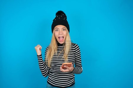 Photo for Positive beautiful kid girl wearing knitted black hat and striped turtleneck over blue background holds modern cell phone connected to headphones, clenches fist from good emotions, exclaims with joy, - Royalty Free Image