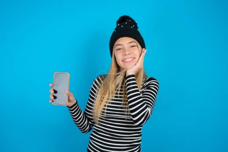 Photo for Beautiful kid girl wearing knitted black hat and striped turtleneck over blue background hold hand modern technology use touch facepalm astonished impressed scream wow omg unbelievable unexpected - Royalty Free Image