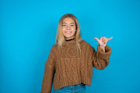 Téléchargez les photos : Blonde enfant fille portant marron pull tricoté sur fond bleu montrant le numéro six Liu avec geste des doigts en langue chinoise signe - en image libre de droit