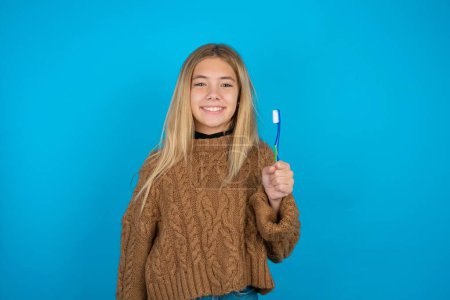 Téléchargez les photos : Gamine blonde portant un pull marron tricoté sur fond bleu tenant une brosse à dents et souriant. Concept de soins dentaires. - en image libre de droit