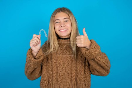 Photo for Blonde kid girl wearing brown knitted sweater over blue background holding an invisible braces aligner and rising thumb up, recommending this new treatment. Dental healthcare concept. - Royalty Free Image