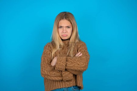 Gloomy dissatisfied blonde kid girl wearing brown knitted sweater over blue background looks with miserable expression at camera from under forehead, makes unhappy grimace