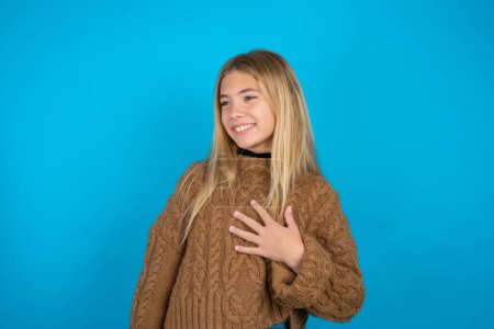 Photo for Joyful blonde kid girl wearing brown knitted sweater over blue background expresses positive emotions recalls something funny keeps hand on chest and giggles happily. - Royalty Free Image
