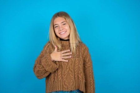 Photo for Blonde kid girl wearing brown knitted sweater over blue background smiles toothily cannot believe eyes expresses good emotions and surprisement - Royalty Free Image