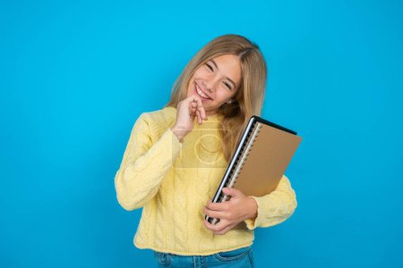 Photo for Beautiful kid girl wearing yellow sweater over blue background laughs happily keeps hand on chin expresses positive emotions smiles broadly has carefree expression - Royalty Free Image
