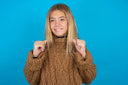 Photo for Blonde kid girl wearing brown knitted sweater over blue background clenches fists and awaits for something nice happened looks away bites lips and waits announcement of results - Royalty Free Image