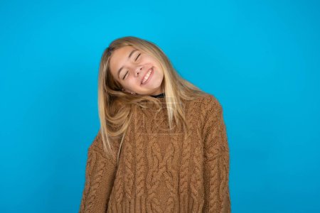 Foto de Chica rubia positiva con suéter de punto marrón sobre fondo azul con expresión de alegría cierra los ojos y se ríe muestra dientes blancos perfectos - Imagen libre de derechos