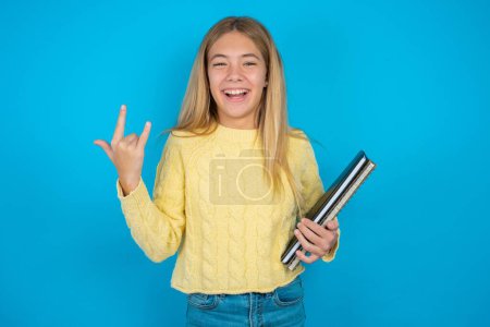 Photo for Beautiful kid girl wearing yellow sweater over blue background makes rock n roll sign looks self confident and cheerful enjoys cool music at party. Body language concept. - Royalty Free Image