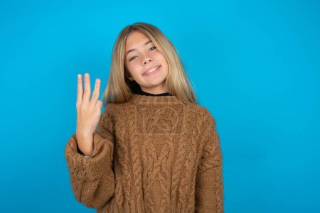 Photo for Blonde kid girl wearing brown knitted sweater over blue background smiling and looking friendly, showing number three or third with hand forward, counting down - Royalty Free Image