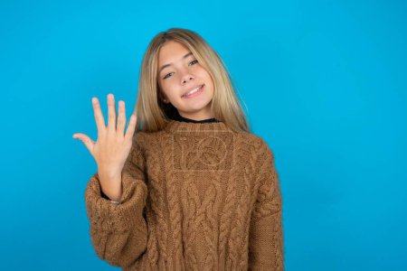 Foto de Niña rubia usando suéter de punto marrón sobre fondo azul sonriendo y mirando amigable, mostrando el número cinco o quinto con la mano hacia adelante, cuenta atrás - Imagen libre de derechos