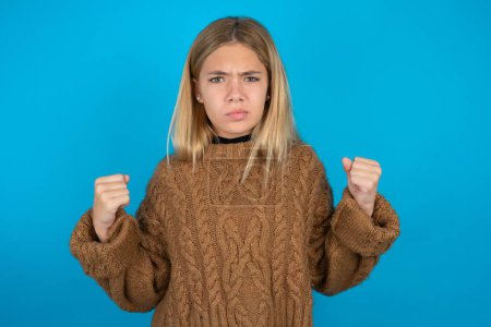 Foto de La niña rubia irritada que usa suéter de punto marrón sobre fondo azul golpea las mejillas con ira y levanta los puños apretados expresa rabia y emociones agresivas. Modelo furioso - Imagen libre de derechos