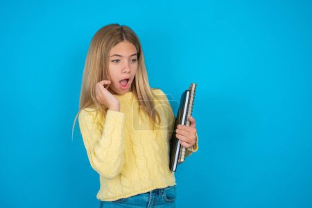 Photo for Astonished beautiful kid girl wearing yellow sweater over blue background looks aside surprisingly with opened mouth. - Royalty Free Image