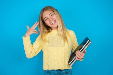 Photo for Beautiful kid girl wearing yellow sweater over blue background foolishness around shoots in temple with fingers makes suicide gesture. Funny model makes finger gun pistol - Royalty Free Image