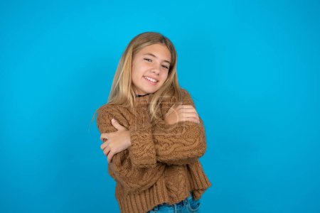 Photo for Charming pleased blonde kid girl wearing brown knitted sweater over blue background embraces own body, pleasantly feels comfortable poses. Tenderness and self esteem concept - Royalty Free Image