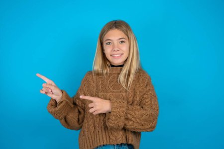Photo for Blonde kid girl wearing brown knitted sweater over blue background points at copy space indicates for advertising gives right direction - Royalty Free Image
