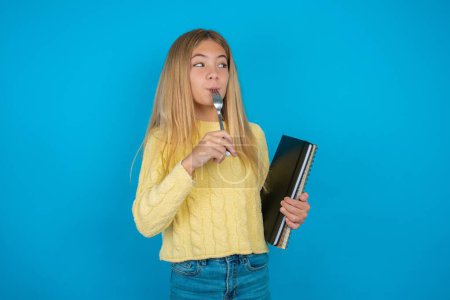 Photo for Photo of dreamy beautiful kid girl wearing yellow sweater over blue background lick fork look empty space - Royalty Free Image