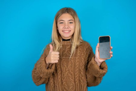 Téléchargez les photos : Blond enfant fille portant brun tricoté pull sur fond bleu Afficher l'écran vide smartphone, pouce vers le haut recommander une nouvelle application - en image libre de droit