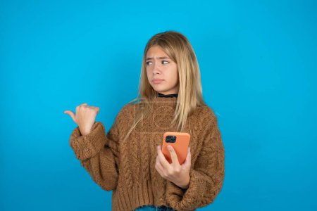 Photo for Blonde kid girl wearing brown knitted sweater over blue background points thumb away and shows blank space aside, holds mobile phone for sending text messages. - Royalty Free Image