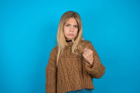 Photo for Blonde kid girl wearing brown knitted sweater over blue background shows fist has annoyed face expression going to revenge or threaten someone makes serious look. I will show you who is boss - Royalty Free Image
