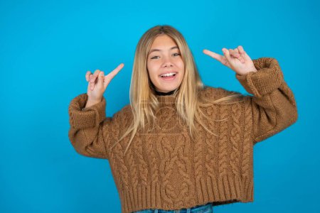 Téléchargez les photos : Joyeux enfant blonde fille portant un pull en tricot brun sur fond bleu démontrant la coiffure - en image libre de droit