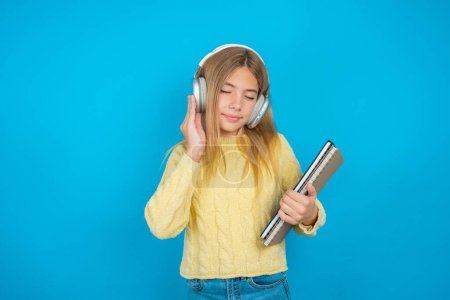 Photo for Pleased beautiful kid girl wearing yellow sweater over blue background enjoys listening pleasant melody keeps hands on stereo headphones closes eyes. Spending free time with music - Royalty Free Image