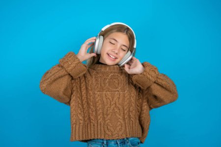 Téléchargez les photos : Blonde enfant fille portant brun tricoté pull sur fond bleu sourit largement se sent très heureux écoute piste de musique préférée via casque sans fil ferme les yeux. - en image libre de droit