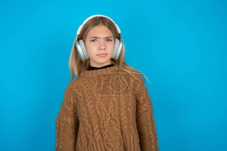 Photo for Displeased blonde kid girl wearing brown knitted sweater over blue background frowns face feels unhappy has some problems. Negative emotions and feelings concept - Royalty Free Image