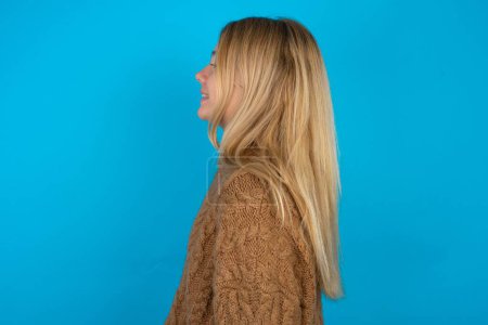 Téléchargez les photos : Profil portrait de belle fille blonde enfant portant chandail tricoté brun sur fond bleu regarder espace vide sourire - en image libre de droit