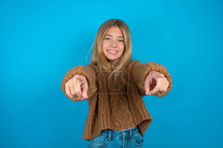 Foto de Rubia niña usando marrón jersey de punto sobre fondo azul alegre y sonriente poiting a cámara - Imagen libre de derechos