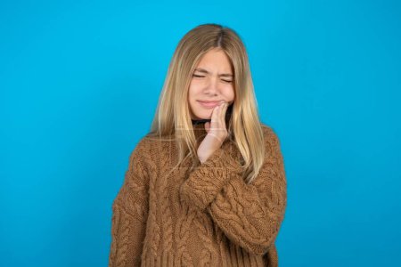 Téléchargez les photos : Enfant blonde fille portant un pull en tricot marron sur fond bleu avec mal de dents - en image libre de droit