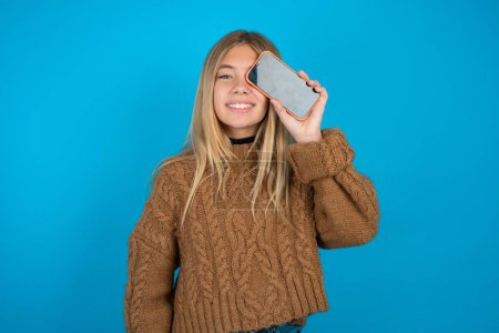 Foto de Niña rubia usando suéter de punto marrón sobre fondo azul sosteniendo teléfono inteligente moderno cubriendo un ojo mientras sonríe - Imagen libre de derechos