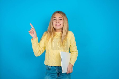 Photo for Beautiful kid girl wearing yellow sweater over blue background holding laptop showing copy empty space ad - Royalty Free Image