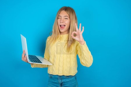 Attraktive fröhlich qualifizierte schöne Kind Mädchen tragen gelben Pullover über blauem Hintergrund mit Laptop zeigt ok-sign winkin