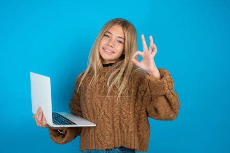 Photo for Positive blonde kid girl wearing brown knitted sweater over blue background hold wireless netbook hand fingers show okey symbol - Royalty Free Image
