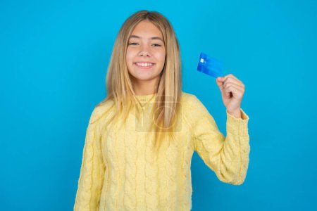 Photo for Photo of happy cheerful smiling positive beautiful kid girl wearing yellow sweater over blue background recommend credit card - Royalty Free Image