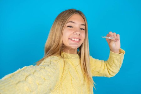 Photo for Beautiful kid girl wearing yellow sweater over blue background make selfie holding an invisible braces aligner, recommending this new treatment. Dental healthcare concept. - Royalty Free Image