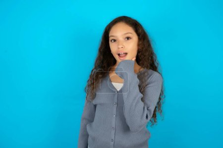 Photo for Optimistic teenager girl wearing grey sweater against blue background keeps hands partly crossed and hand under chin, looks at camera with pleasure. - Royalty Free Image