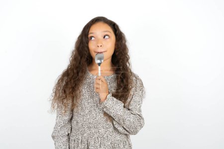 Foto de Muchacha adolescente muy hambrienta con vestido gris sobre fondo blanco sosteniendo cuchara en la boca sueño de comida sabrosa - Imagen libre de derechos