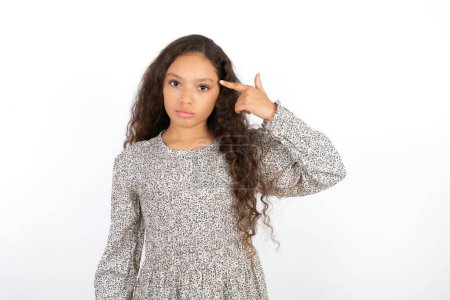 Photo for Unhappy Teenager girl wearing grey dress against white background makes suicide gesture and imitates gun with hand, curves lips keeps two fingers on temple, shoots, being tired of everything, - Royalty Free Image