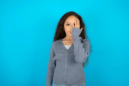 Téléchargez les photos : Adolescent fille portant pull gris sur fond bleu couvrant un oeil avec sa main, sourire confiant sur le visage et l'émotion surprise. - en image libre de droit