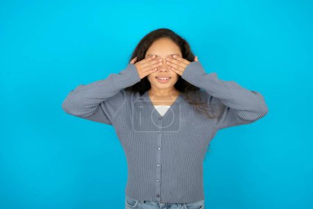 Téléchargez les photos : Adolescente portant un pull gris sur fond bleu couvrant les yeux avec les mains souriantes gaies et drôles. Concept aveugle. - en image libre de droit