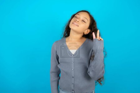 Foto de Adolescente usando suéter gris sobre fondo azul sonriendo con la cara feliz guiñando el ojo a la cámara haciendo signo de victoria. Número dos.. - Imagen libre de derechos