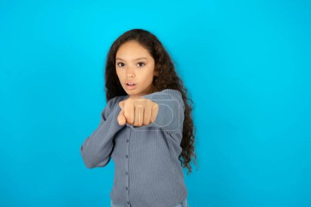 Photo for Teenager girl wearing grey sweater against blue background Punching fist to fight, aggressive and angry attack, threat and violence - Royalty Free Image