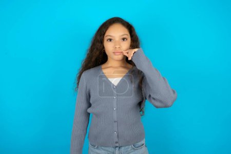 Foto de Adolescente chica usando suéter gris contra fondo azul boca y labios cerrados como cremallera con los dedos. Secreto y silencioso, tabú hablando. - Imagen libre de derechos