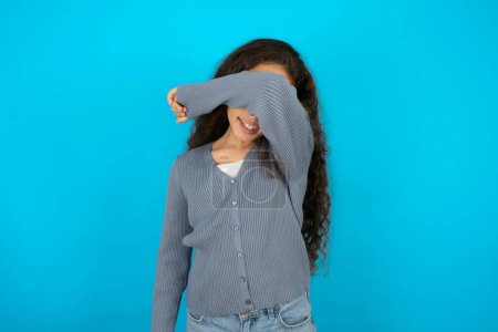 Photo for Teenager girl wearing grey sweater against blue background covering eyes with arm smiling cheerful and funny. Blind concept. - Royalty Free Image