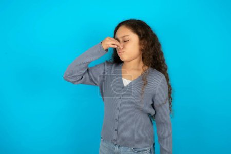 Téléchargez les photos : Adolescente portant un pull gris sur fond bleu sentant quelque chose de puant et dégoûtant, odeur intolérable, retenant la respiration avec les doigts sur le nez. Mauvaise odeur - en image libre de droit