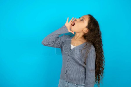 Téléchargez les photos : Adolescente portant un pull gris sur fond bleu criant et criant haut et fort à côté avec la main sur la bouche. Concept de communication. - en image libre de droit