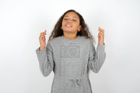 Photo for Teenager girl wearing grey dress against white background gesturing finger crossed smiling with hope and eyes closed. Luck and superstitious concept. - Royalty Free Image